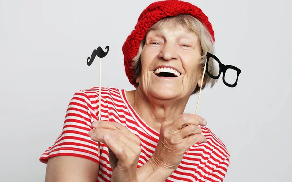 Engraçada avó com bigode falso e óculos, ri e se prepara para a festa sobre fundo cinza — Fotografia de Stock