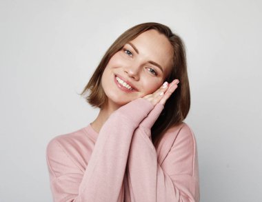 Tatlı gülüşü olan güzel genç bir kadın açık gri arka planda yalnız poz veriyor..