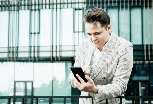 Jovem empresário vestido com roupas formais em pé fora de um edifício de vidro e usando telefone celular — Fotografia de Stock