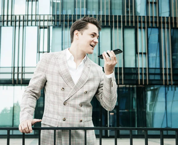 Jovem empresário vestido com roupas formais em pé fora de um edifício de vidro e usando telefone celular — Fotografia de Stock