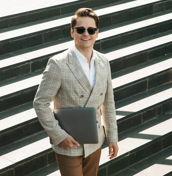 Guapo joven hombre de negocios sonriente pasar tiempo al aire libre en la ciudad, utilizando el ordenador portátil en las escaleras —  Fotos de Stock