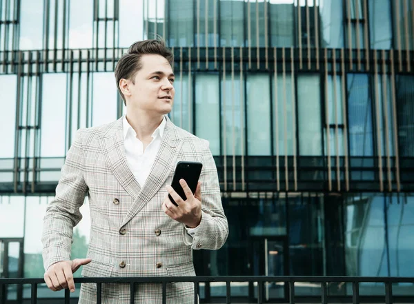 Joven hombre de negocios vestido con ropa formal de pie fuera de un edificio de vidrio y utilizando el teléfono móvil —  Fotos de Stock