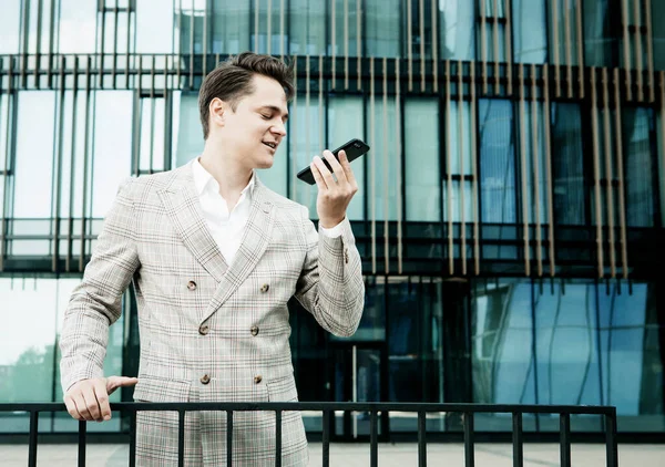 Молодой бизнесмен в формальной одежде, стоящий у стеклянного здания и пользующийся мобильным телефоном — стоковое фото