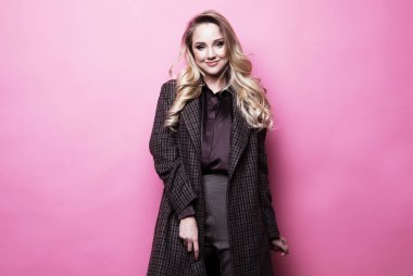 Güzel bir kadın. Uzun sarı kıvırcık saçlı. Pembe arka planda rahat giyiniyor.