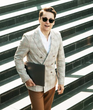 Yakışıklı, gülümseyen genç iş adamı şehirde vakit geçiriyor, merdivenlerde dizüstü bilgisayar kullanıyor.