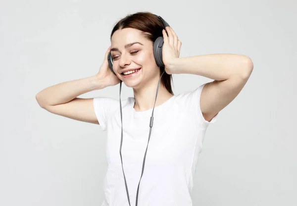 Mujer joven con auriculares escuchando música de pie sobre un fondo gris en camiseta blanca —  Fotos de Stock