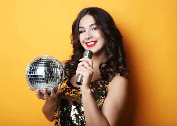 Woman in evening dress holding microphone and disco ball over yellow background — Stock Photo, Image