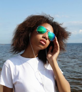 Güneş gözlüklü, kulaklıklı, cep telefonuyla müzik dinleyen büyüleyici afro-amerikalı genç kadın. Sıradan giyinmiş. Denizin kenarında eğlenmek için..