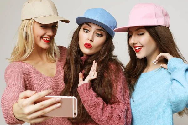 Trois bonnes copines heureuses portant des pulls et des casquettes faisant du selfie sur smartphone. — Photo