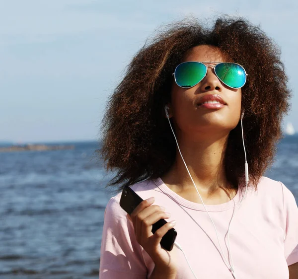 Urocza, niesamowita afro-amerykańska młoda kobieta w okularach przeciwsłonecznych słuchająca muzyki w słuchawkach na telefonie komórkowym. Ubrany swobodnie. — Zdjęcie stockowe