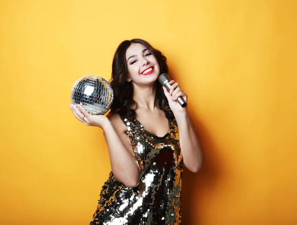 Woman in evening dress holding microphone and disco ball over yellow background — Stock Photo, Image