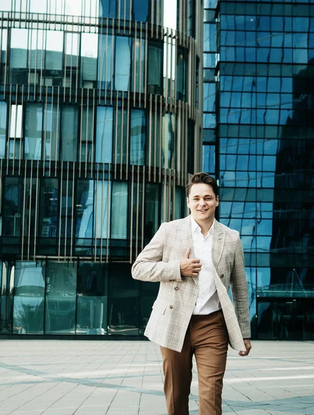 Joven hombre feliz en un traje de negocios posa cerca de un moderno centro de negocios en un día de verano —  Fotos de Stock