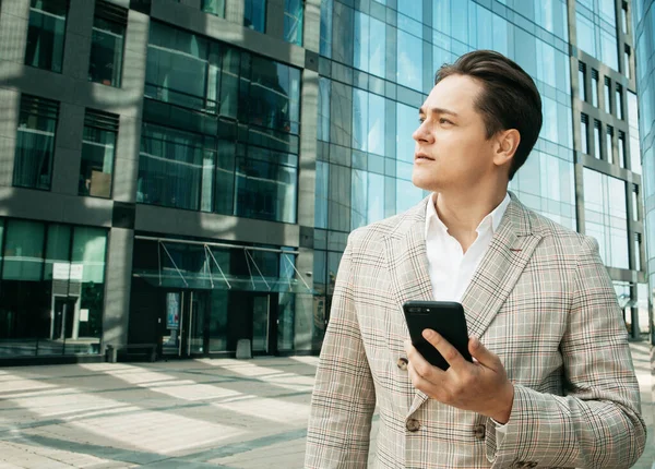 Junger Geschäftsmann in formeller Kleidung steht vor einem gläsernen Gebäude und benutzt Mobiltelefone — Stockfoto