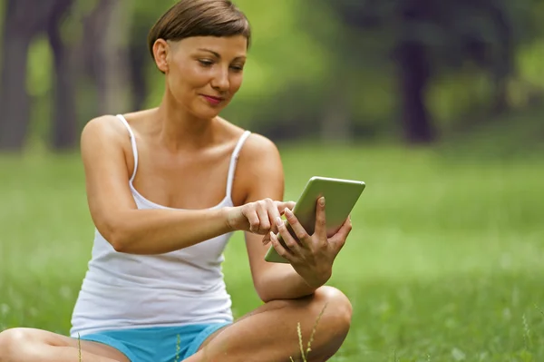 Jovem mulher usando tablet ao ar livre localização na grama, sorrindo . — Fotografia de Stock