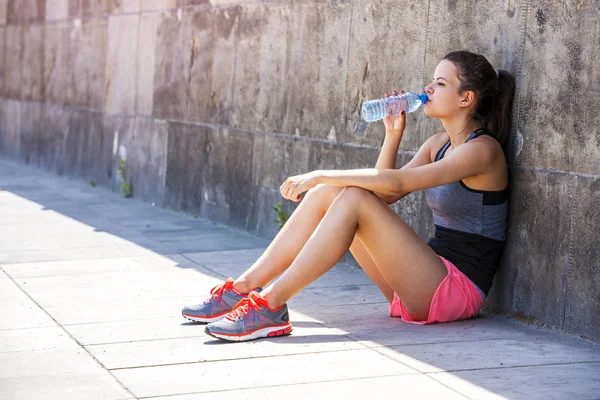 Thirsty female jogger drinking fresh water and siting after trai — Stock Photo, Image