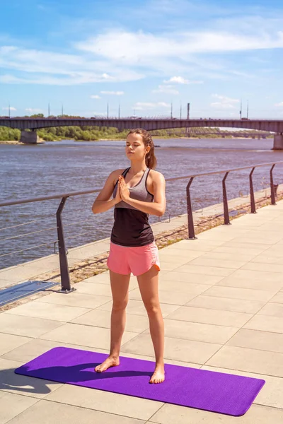 Young and sporty woman doing yoga standing on mat by the river i — Stock Photo, Image