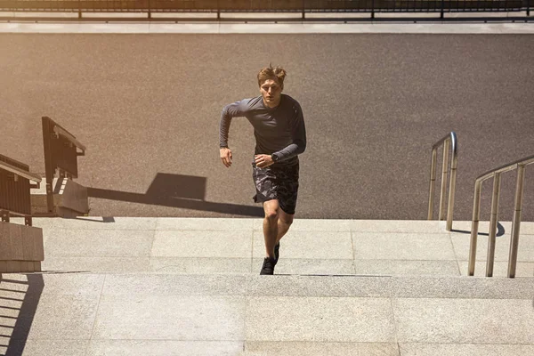 Male athlete running up a flight of stairs with speed, sporty young man in sporty t-shirt training or working out outdoors while jogging up the steps, flare sun