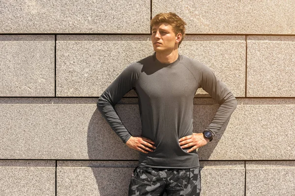 Muscular build young runner, tired after training outdoors, athletic jogger in bright sportswear resting after run on beautiful concrete wall background, fitness concept