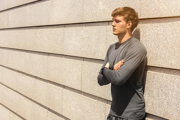 Muscular build young runner crossed arms, tired after training outdoors, athletic jogger in bright sportswear resting after run on beautiful concrete wall background, fitness concept