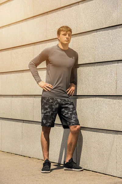 Muscular build young runner, tired after training outdoors, athletic jogger in bright sportswear resting after run on beautiful concrete wall background, fitness concept