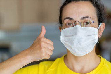 Koruyucu steril tıbbi maskeli genç İspanyol kadın evinde kameraya bakıyor. Göster, tamam. Elle.