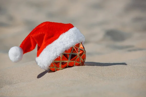 Cappello rosso di Babbo Natale indossato sulla palla di Natale sdraiato sulla spiaggia, sulla sabbia. Vacanze di anno nuovo nell'oceano o nel mare . — Foto Stock