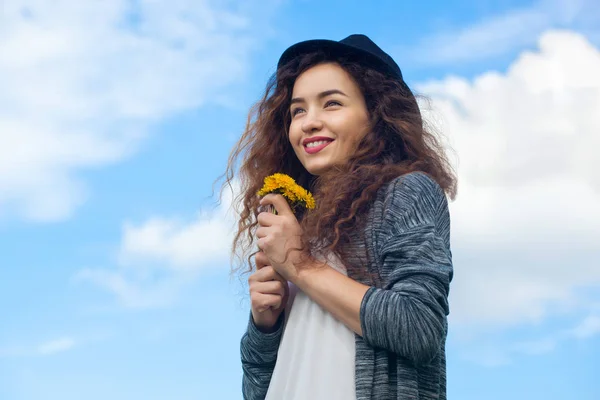 Menina atraente, jovem em jeans e um chapéu preto segurando um dente-de-leão florescendo — Fotografia de Stock