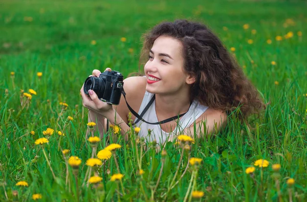 Linda, atraente menina-fotógrafo com cabelo encaracolado segura uma câmera e deitado na grama com dentes-de-leão florescendo na primavera ao ar livre — Fotografia de Stock