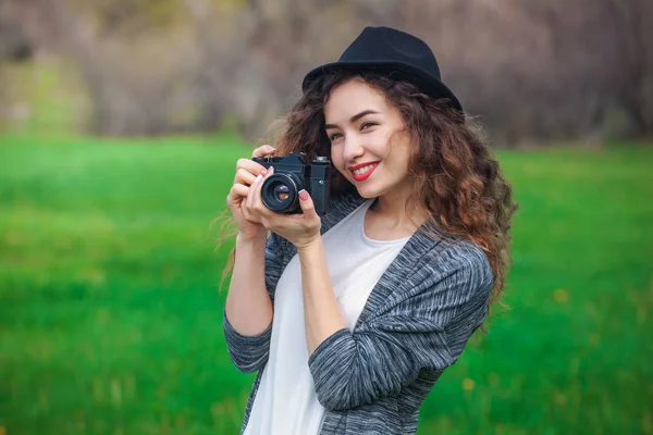 Menina-fotógrafo bonita com cabelo encaracolado segura uma câmera e fazer uma foto, primavera ao ar livre no parque . — Fotografia de Stock