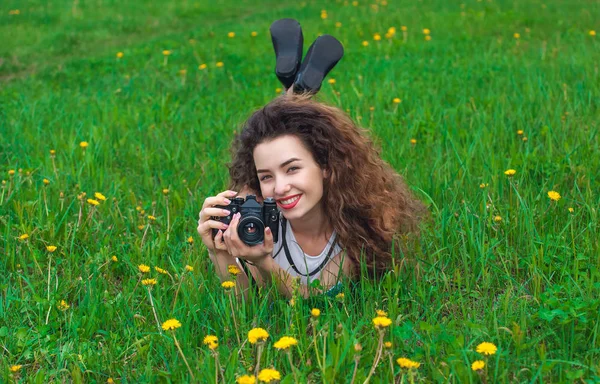 Linda, atraente menina-fotógrafo com cabelo encaracolado segura uma câmera e deitado na grama com dentes-de-leão florescendo na primavera ao ar livre — Fotografia de Stock