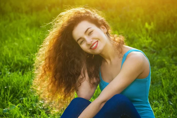 Menina atraente, jovem com cabelo encaracolado sentado na grama verde no gramado e sorrindo para o fotógrafo — Fotografia de Stock