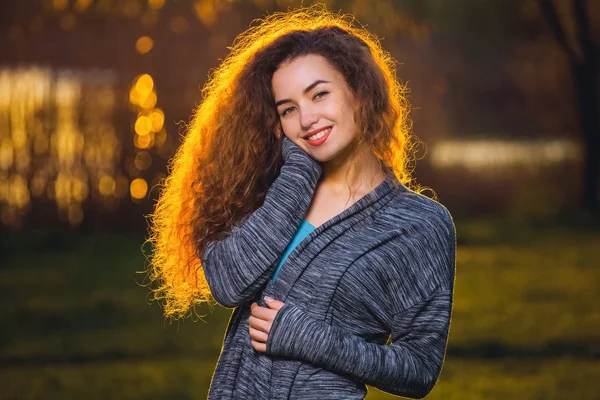 Sorrindo menina bonita com encaracolado, cabelo iluminado pelo sol em um belo fundo — Fotografia de Stock