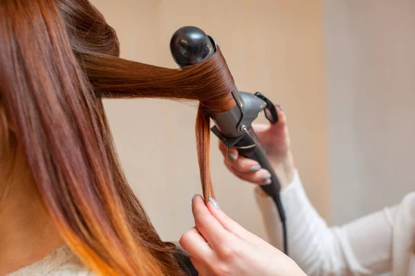 Парикмахер делает прическу девушки с длинными рыжими волосами в салоне красоты. Создание кудрей с щипцами для завивки . — стоковое фото