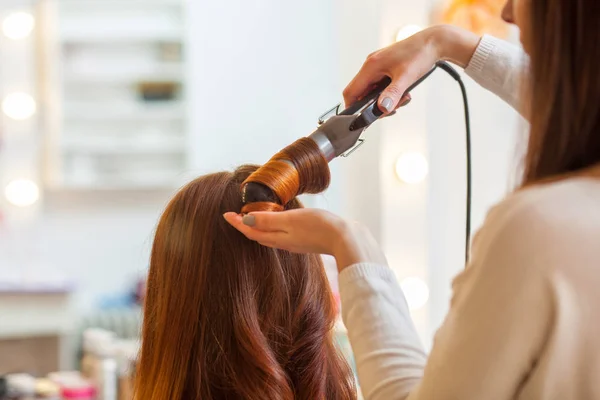 Cabeleireiro faz penteado menina com cabelo longo vermelho em um salão de beleza. Criar cachos com ferros de ondulação . — Fotografia de Stock