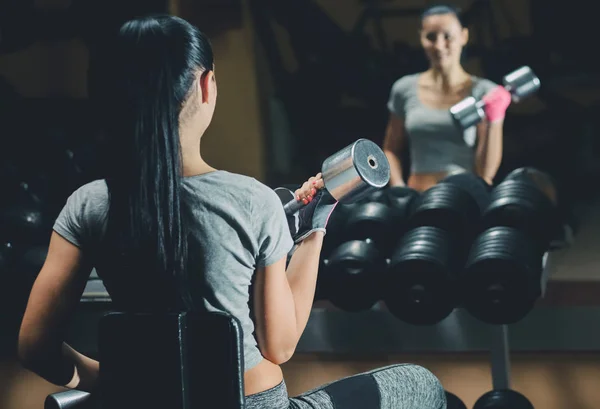 Slim bodybuilder girl lifts heavy dumbbell standing in front of the mirror while training in the gym. — Stock Photo, Image