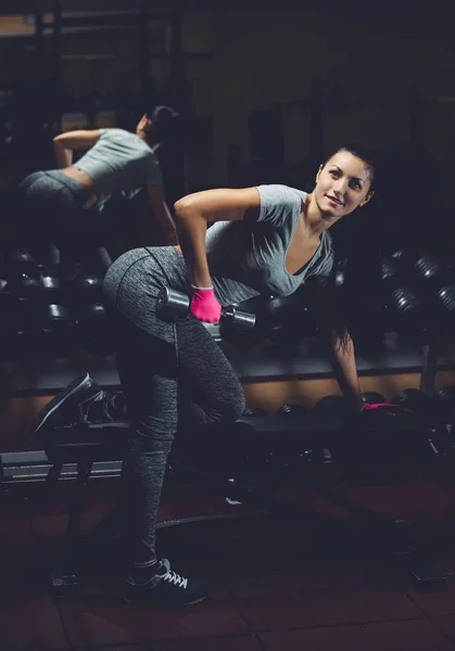 Slim, bodybuilder girl, lifts heavy dumbbell standing in front of the mirror while training in the gym.