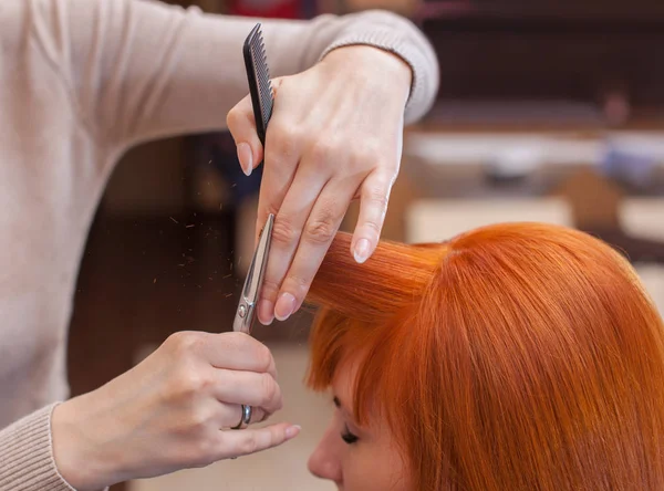 O cabeleireiro faz um corte de cabelo com tesoura de cabelo para um jovem com cabelo vermelho menina — Fotografia de Stock