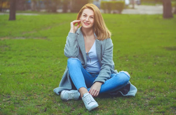 Ελκυστική, νεαρή ξανθιά κοπέλα σε ένα γκρι παλτό και τζιν που κάθεται στο γρασίδι — Φωτογραφία Αρχείου