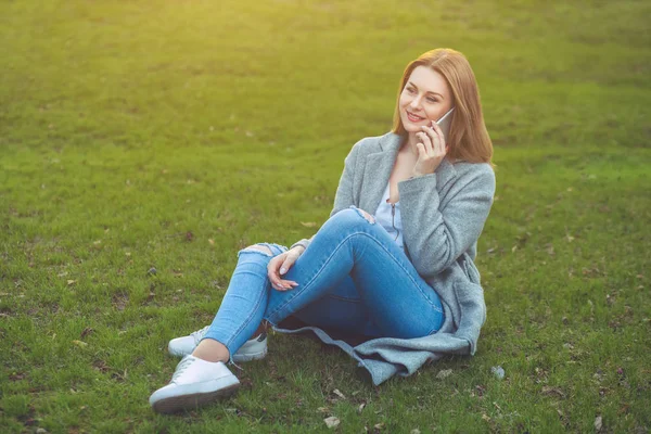 Vonzó szőke lány ül a fűben mobiltelefonon beszél — Stock Fotó