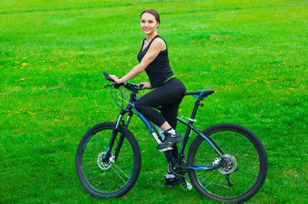 Jovem mulher atraente montando uma bicicleta de montanha em um parque em um gramado verde — Fotografia de Stock