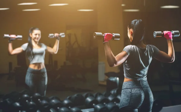 Slim, bodybuilder girl, lifts heavy dumbbell standing in front of the mirror while training in the gym. — Stock Photo, Image