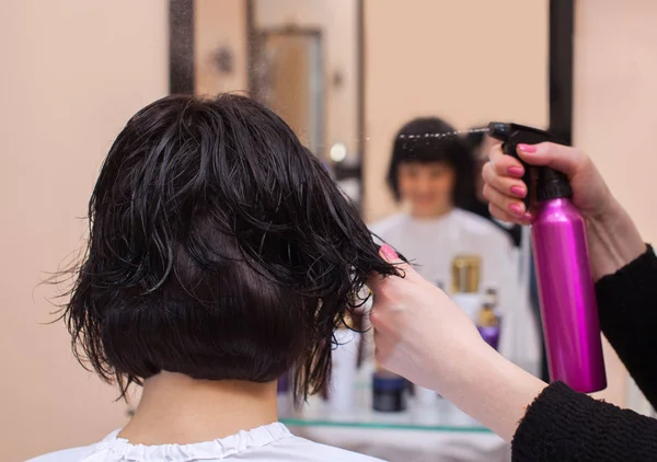 Парикмахер перед сушкой волос брызгает спреем на волосы брюнетки — стоковое фото