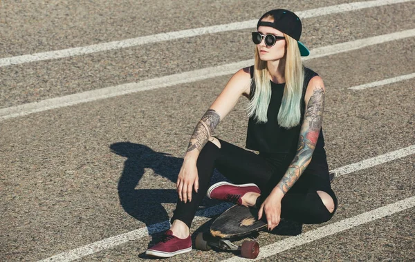 A beautiful, sexy hipster blonde with blue hair in tattoo sits on a longboard  with glasses — Stock Photo, Image