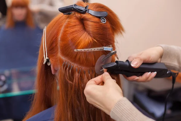 O cabeleireiro faz extensões de cabelo para uma jovem, menina de cabelos vermelhos — Fotografia de Stock