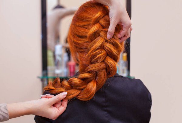 Beautiful, red-haired girl with long hair, hairdresser weaves a French braid