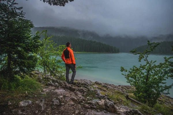a man stands by a mountain lake in cloudy weather in the mountains in summer with slopes and green trees