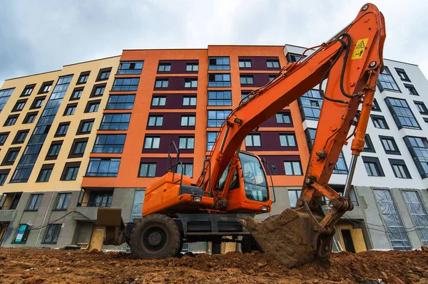 orange excavator digs soil against the background of a new bright house