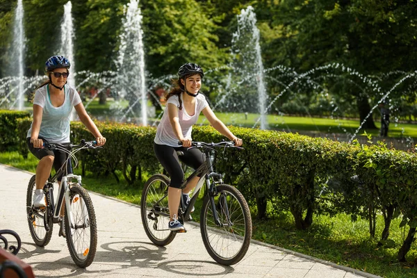 Dos Mujeres Cascos Montando Bicicletas Parque Verano Con Fuentes Fondo —  Fotos de Stock