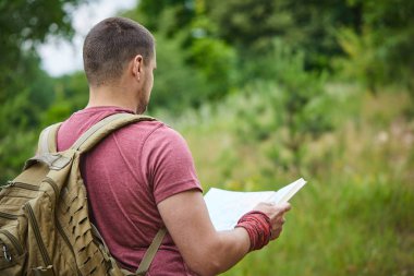 Güneşli bir günde çayırda yürüyen haritalı ve sırt çantalı genç bir adam.