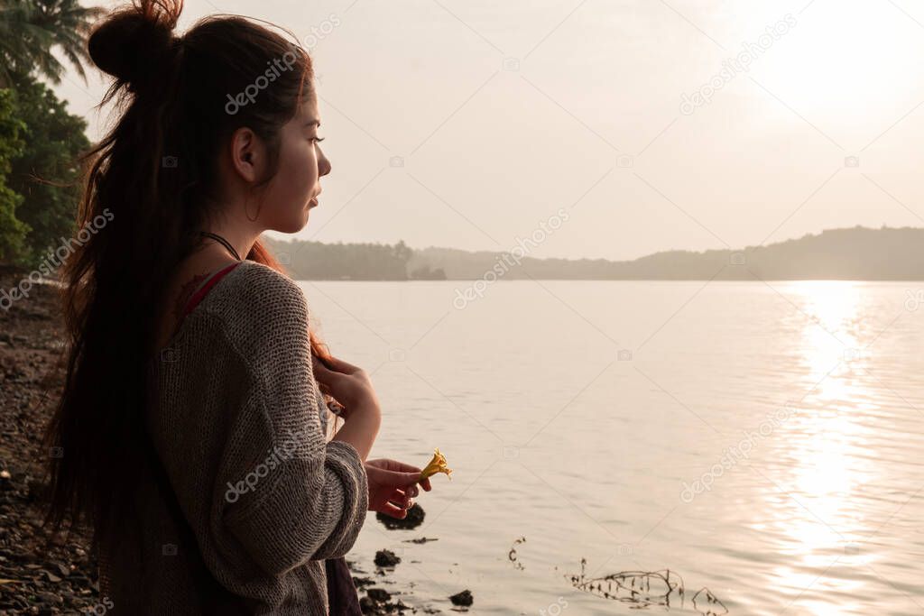 Asian girl stands on the seashore with a flower in her hands, meets the sunset